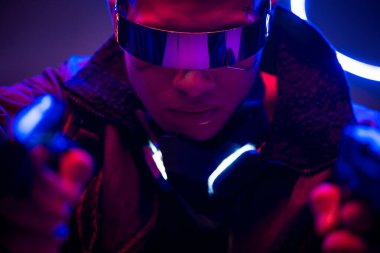 Fütürist gözlüklü silahlı çift ırklı siber punk oyuncusunun seçici odak noktası neon ışıklarının yanında silah tutması. 