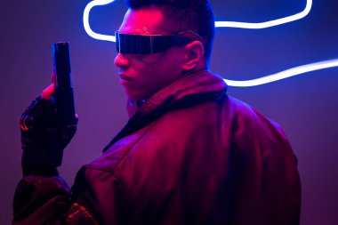 Melez ırk siber punk oyuncusunun fütüristik gözlüklü yan görüntüsü neon ışıklarının yanında silah taşıyor. 