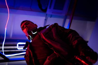 low angle view of mixed race cyberpunk player standing near blue neon lighting  clipart