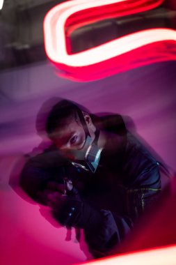 Kırmızı neon ışığın yanında maskeli silahlı, çift ırklı siber punk oyuncunun hareket bulanıklığı. 