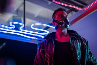 Maskeli karışık ırk siber punk oyuncusu neon ışığın yanında duruyor ve başka tarafa bakıyor.