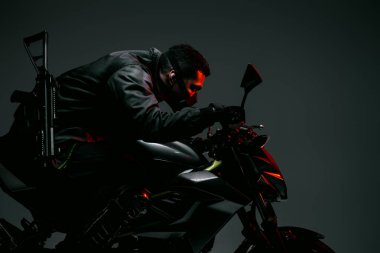 profile of armed bi-racial cyberpunk player in mask riding motorcycle on grey  clipart