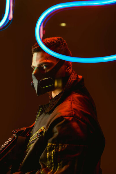 селективный фокус опасного бирасового киберпанка в маске, держащего пистолет рядом с синим неоновым светом на черном
 