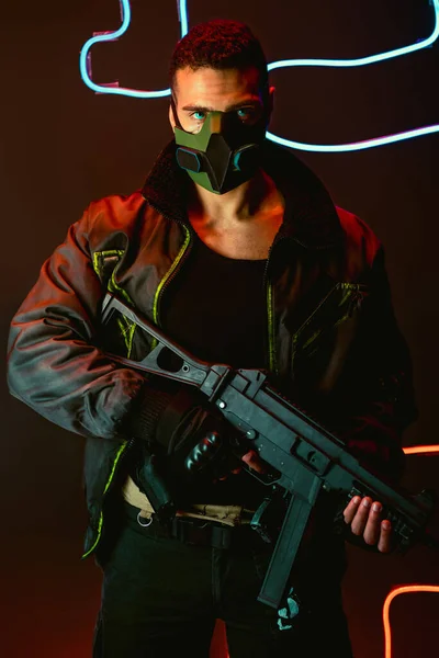 bi-racial cyberpunk player in protective mask with gun near neon lighting on black