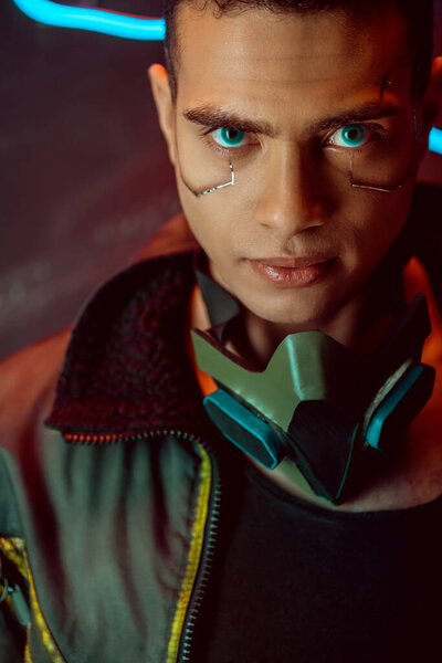 handsome and bi-racial cyberpunk player looking at camera near neon lighting on black 