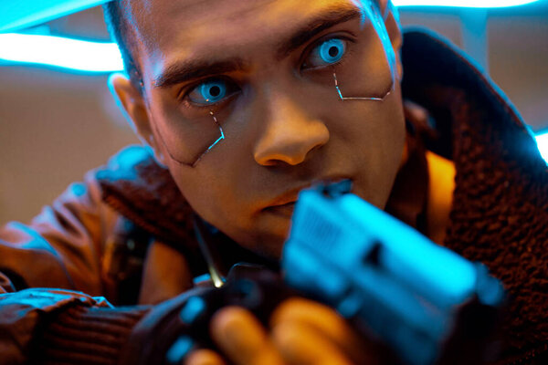 селективный фокус киберпанка смешанной расы с металлическими пластинами на лице и голубыми глазами, держащими пистолет возле неонового освещения
 