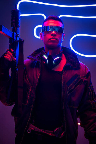 dangerous mixed race cyberpunk player in futuristic glasses holding gun near neon lighting 