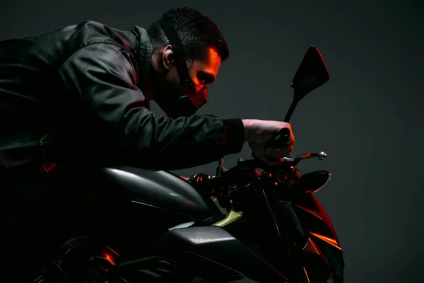 profile of mixed race cyberpunk player in mask riding motorcycle on grey