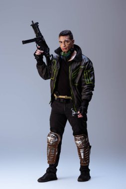 Yakışıklı melez siber punk oyuncusu gri üzerinde dururken silah tutuyor.  