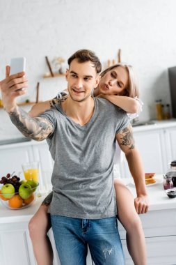 Mutfakta dövmeli erkek arkadaşının yanında selfie çekerken yüzünü buruşturan kadınların seçici odağı. 