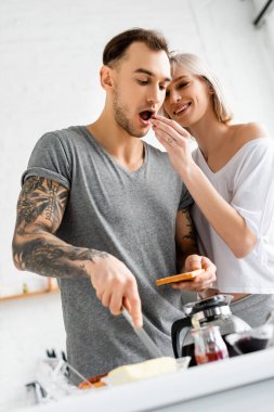 Selective focus of smiling woman feeding tattooed boyfriend with grape during breakfast in kitchen  clipart