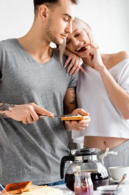 Selective focus of tattooed man with toast and knife looking at smiling girlfriend eating grape near breakfast in kitchen  clipart