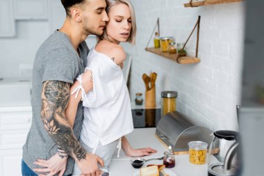 Side view of tattooed man touching beautiful blonde girlfriend near breakfast in kitchen  clipart
