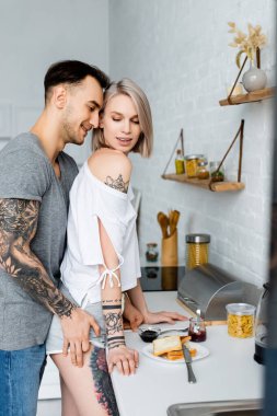 Side view of tattooed man touching hip of sensual blonde girl near breakfast on kitchen worktop  clipart