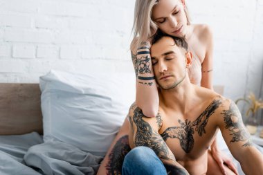 Sütyenli seksi kadın yatakta üstsüz dövmeli erkek arkadaşını kucaklıyor. 