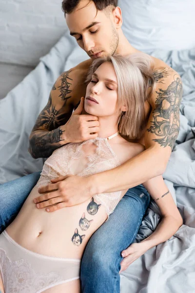 Handsome tattooed man embracing and touching face of sensual girlfriend in underwear on bed