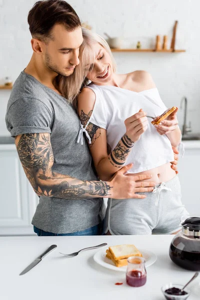 英俊的纹身男子在咖啡壶边和厨房桌子上的果酱旁边拥抱着笑着的女朋友 举杯庆祝 — 图库照片