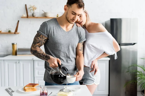 Beautiful Girl Embracing Tattooed Boyfriend Pouring Coffee Kitchen — Stock Photo, Image