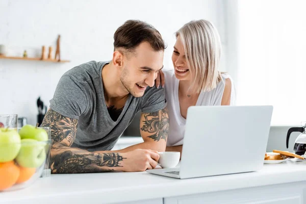 微笑的女人在笔记本电脑旁边拥抱纹身的男朋友 在厨房桌子上拥抱咖啡 — 图库照片