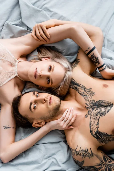 Top view of tattooed couple looking at camera while touching each other on bed