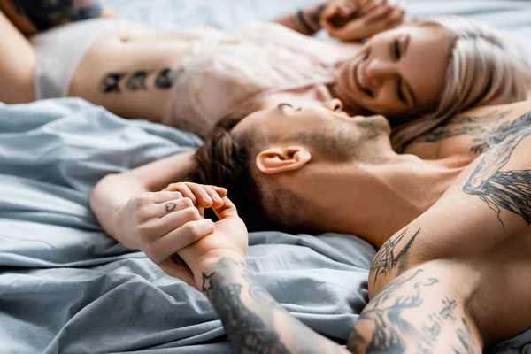 Selective focus of muscular tattooed man smiling at beautiful girlfriend in lingerie on bed