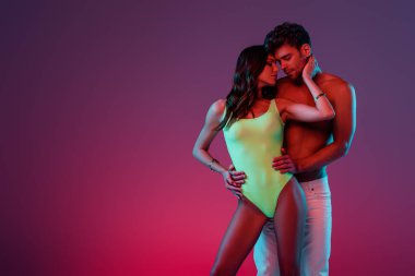 Seksi gömleksiz adam, mayolu güzel kızı kucaklıyor mor arka planda boynuna dokunuyor.