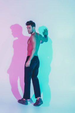 Koyu mavi kotlu seksi gömleksiz adam duvara dokunuyor ve arka plandaki mavi ve mor gölgeli kameraya bakıyor.