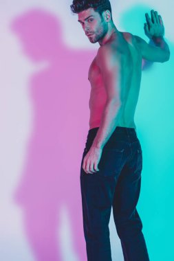 Koyu mavi kotlu seksi gömleksiz adam arka planda mavi ve mor gölgelerle dikilirken duvara dokunuyor.