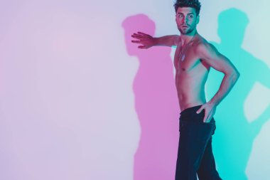 Koyu mavi kotlu seksi gömleksiz adam duvara dokunuyor, el ele tutuşuyor ve mavi ve mor gölgelerle arka plana bakıyor.
