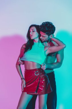 Seksi adam tutkulu bir kıza dokunuyor mavi arka planda boynunu sarıp turkuaz ve mor gölgelerle sarılıyor.