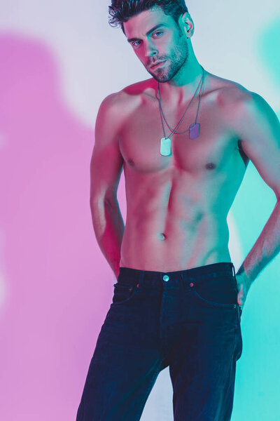 sexy, shirtless man in dark blue jeans holding hands behind back and looking at camera on background with violet shadow