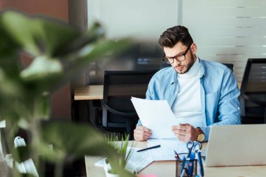 Selective focus of concentrated IT worker looking at papers at table in coworking space clipart