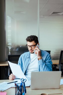 Bilgi işlem işçisi elinde kağıtlarla birlikte çalışma alanındaki masada akıllı telefondan konuşuyor.