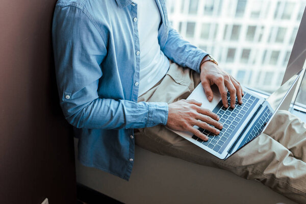 Cropped view of IT worker using laptop on windowsill near window