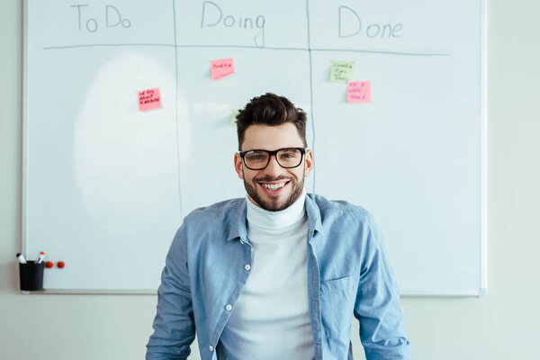 Scrum master looking at camera and smiling near white board with stickers and spreadsheet with to do, doing, done lettering 