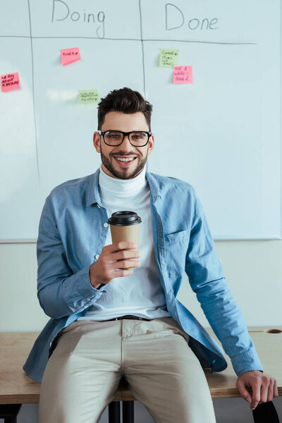 Scrum master looking at camera and smiling with paper cup of coffee near white board with spreadsheet with doing, done lettering and stickers 