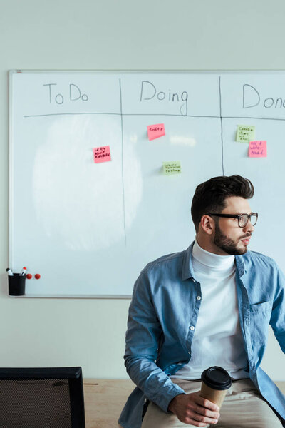 Scrum master looking away with paper cup of coffee near white board with spreadsheet with to do, doing, done lettering and stickers 