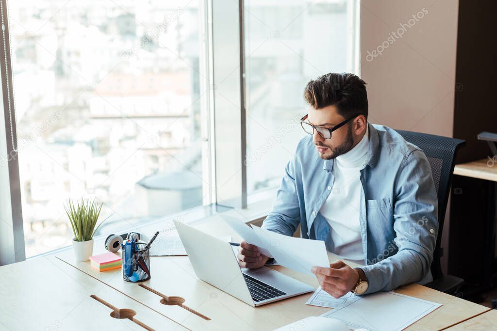 High angle view of concentrated IT worker looking at papers near laptop at table in coworking space