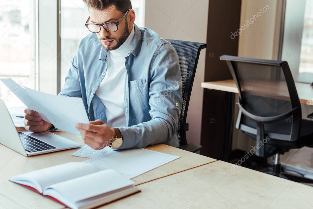 Concentrated IT worker looking at papers near laptop at table in coworking space