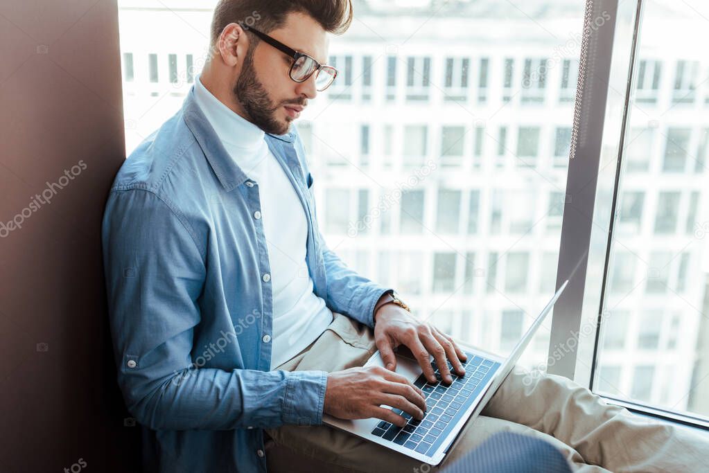 Selective focus of IT worker using laptop on windowsill near windows