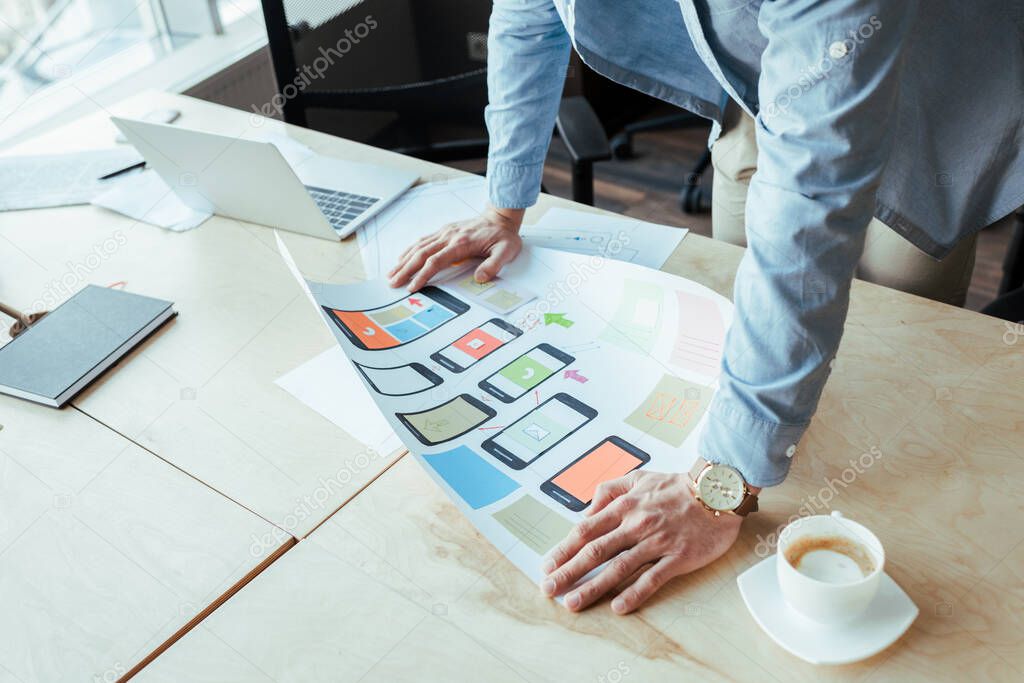 Cropped view of UI designer with wove paper near cup of coffee at table in coworking space