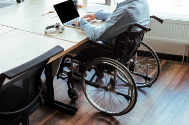 Tekerlekli sandalyedeki engelli bilişim işçisinin kulaklıkların yanında dizüstü bilgisayarla çalışırken görüntüsü kesilmiş. 