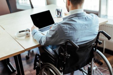 Cropped view of disabled IT worker on wheelchair working with laptop near headphones at table in coworking space clipart