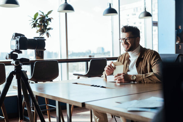 Selective focus of blogger in front of digital camera smiling and holding opened box at table in coworking space
