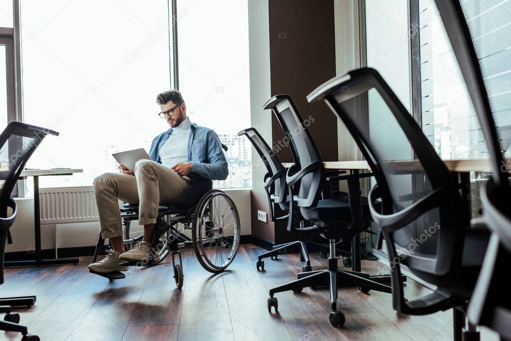 Selective focus of disabled IT worker on wheelchair working with laptop near windows in coworking space
