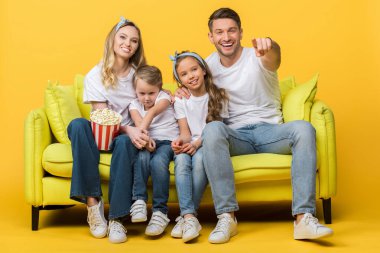 Gülümseyen ebeveynler ve çocuklar kanepede sarı mısır kovası ile film izleyip işaret ediyorlar. 