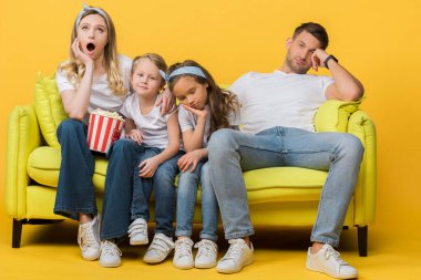 Sıkılmış ebeveynler ve çocuklar kanepede film izlerken ellerinde patlamış mısır kovası ve sarı 