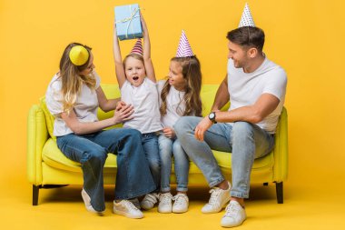 Mutlu bir aile, doğum günü partisi şapkaları içinde, heyecanlı bir oğul, sarı koltukta bir hediye kutusu tutuyor. 