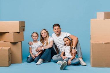Mutlu ebeveynler çocukları kucaklıyor ve taşınmak için karton kutularla mavinin üzerinde oturuyorlar.