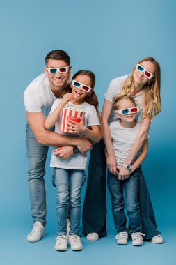 Mutlu ebeveynler ve çocuklar 3 boyutlu gözlüklerle film izliyor ve mavi üzerinde patlamış mısır kovası tutuyorlar.